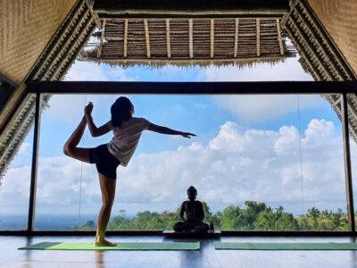 Young woman doing balance pose while practicing yoga near a Buddha statue with blue sky view background at morning time