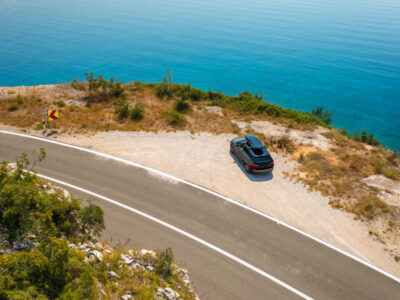 Car with a roof box, parked on the edge of a cliffside road, with the azure sea below reflecting the clear blue sky.