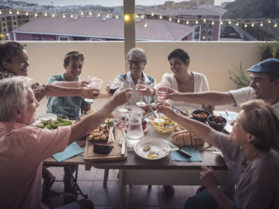 Group of mixed ages friends toasting with glasses having dinner in the evening at home in outdoor terrace with city view - mixed generations have fun all together clinking and enjoying leisure time dining
