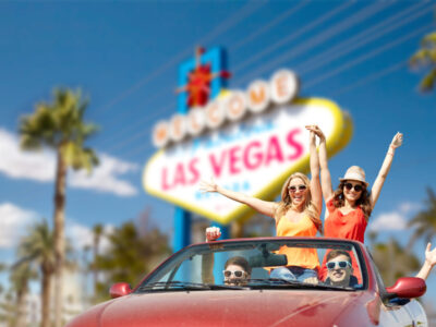 summer holidays, road trip and travel concept - happy friends driving in convertible car and waving hands over welcome to fabulous las vegas sign background