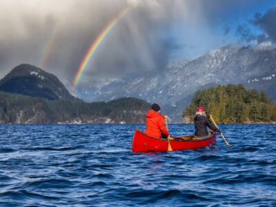 Couple friends on a wooden canoe are paddling in water. Dramatic Rainbow Sky Art Render. Taken in Indian Arm, near Deep Cove, North Vancouver, British Columbia, Canada. Concept: Adventure, Sport