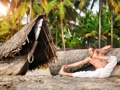 Yoga aakarna dhanurasana archer pose by fit man in white trousers on the beach near the fishermen boats in India